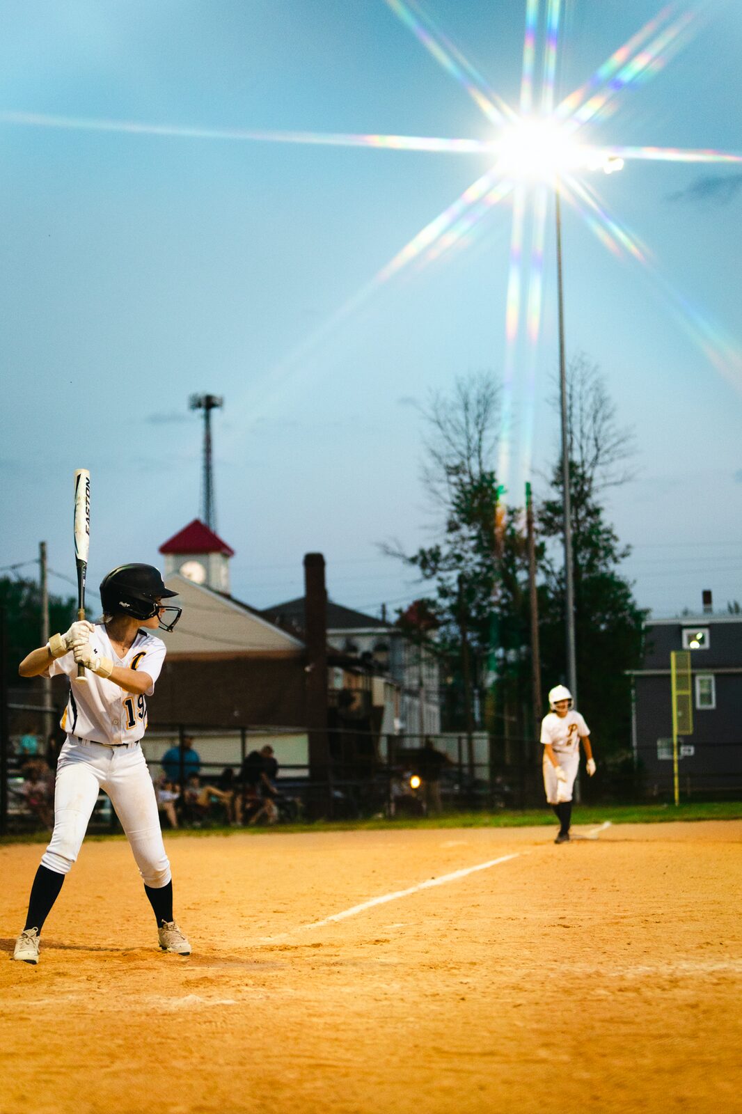 Softball batter set in the box at dusk with a stadium light bursting into a starburst above the field