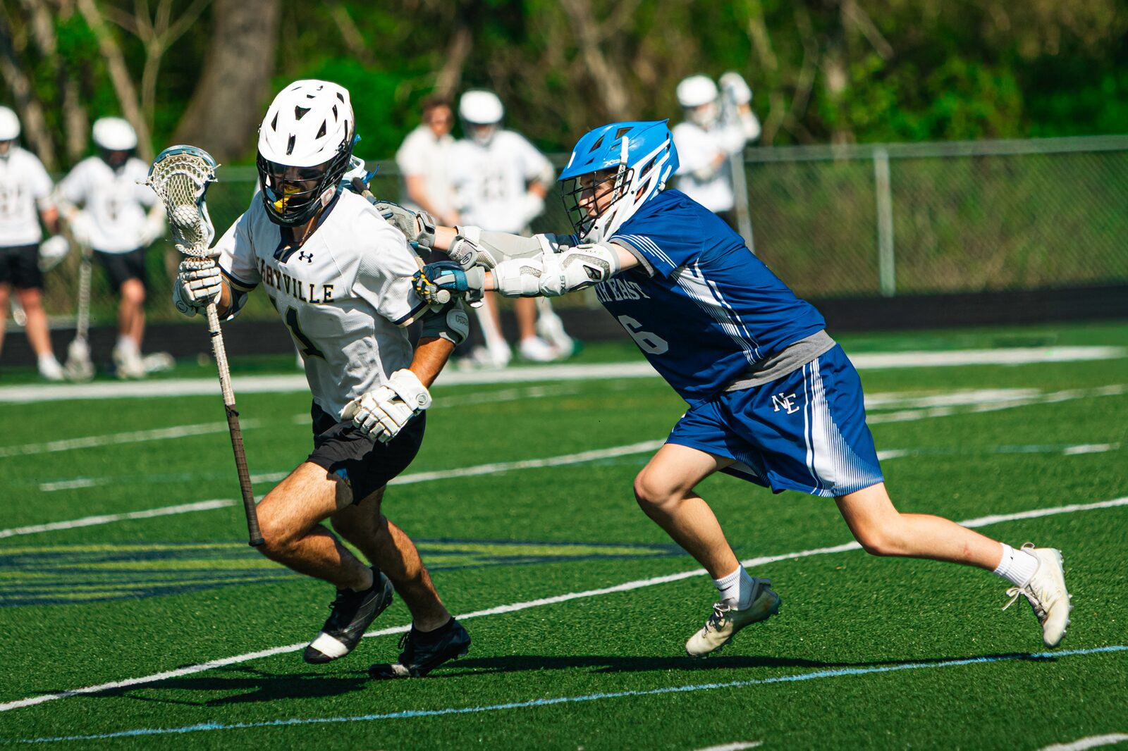 Lacrosse defender throwing a check on a ball carrier at midfield during a day game