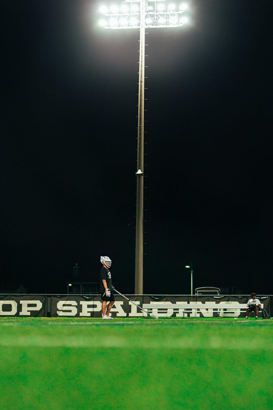 Lone lacrosse player standing on an empty green field beneath a tall stadium light at night