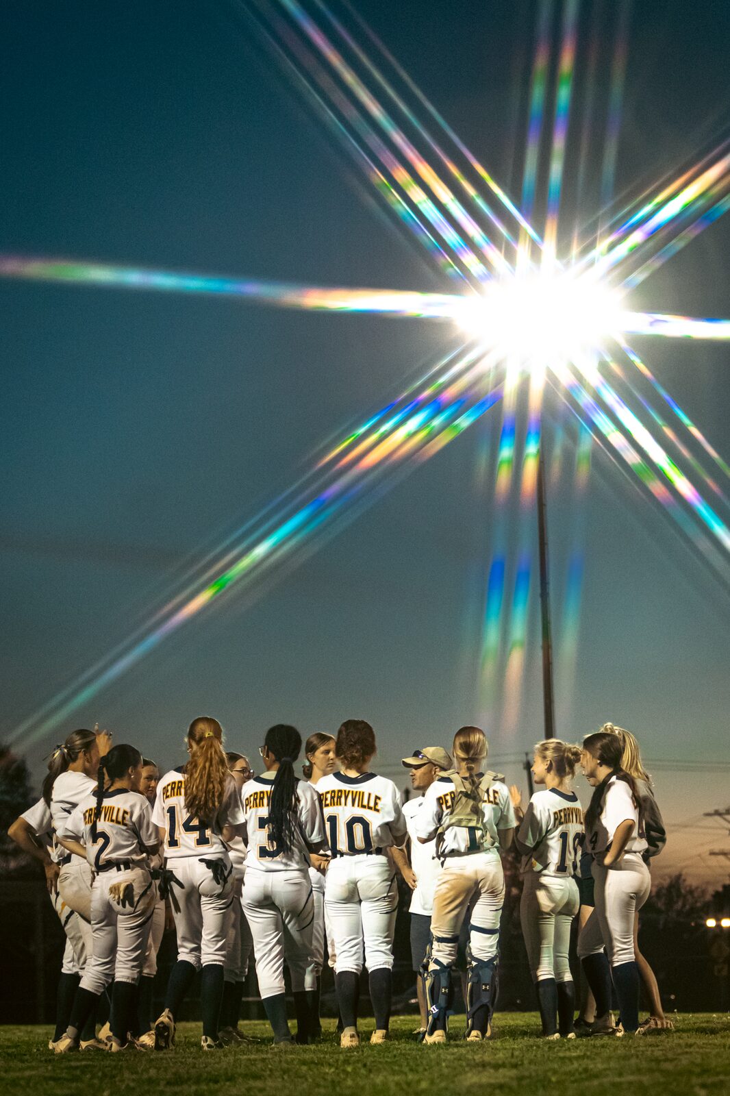Softball team gathered in a huddle at dusk under the field lights