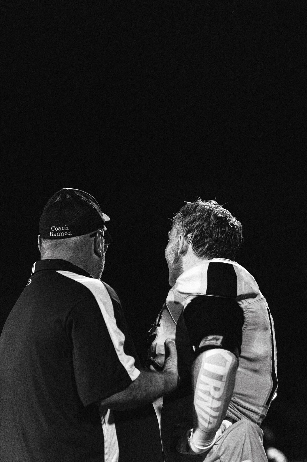 Black and white photo of a coach leaning in to talk with a player on the sideline at night