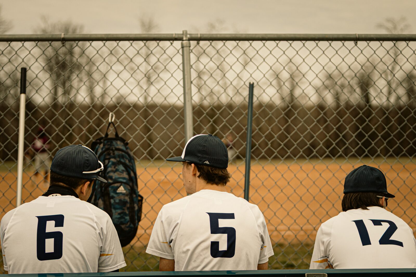 Three teammates sitting on the bench behind the backstop fence watching the game