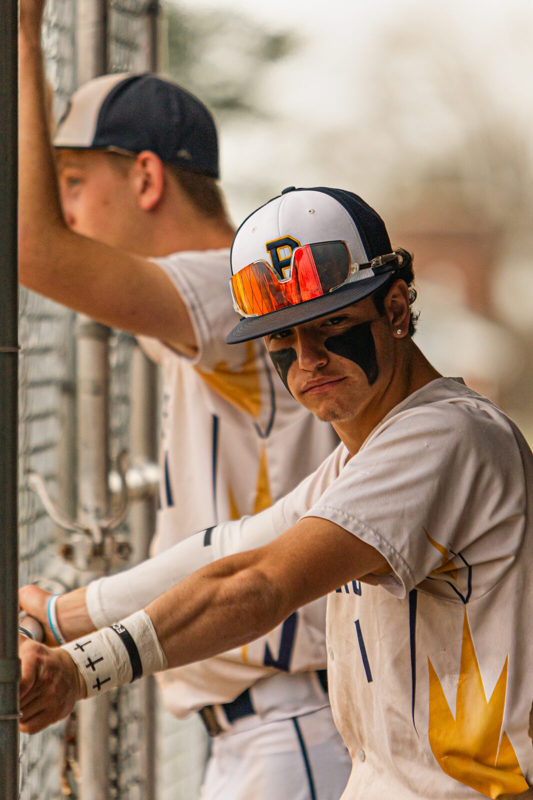 Player in sunglasses and eye black leaning on the dugout fence looking out at the field