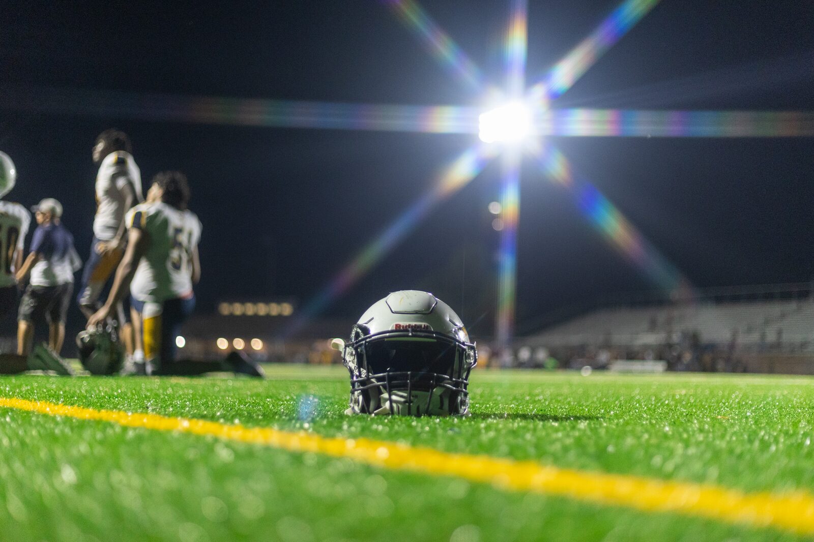A football helmet resting on the turf beside a yard line as a stadium light bursts into a starburst overhead