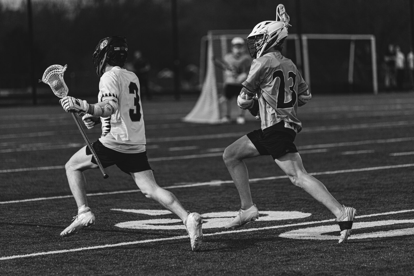 Black and white photo of two lacrosse players at full stride chasing a loose ball up the field