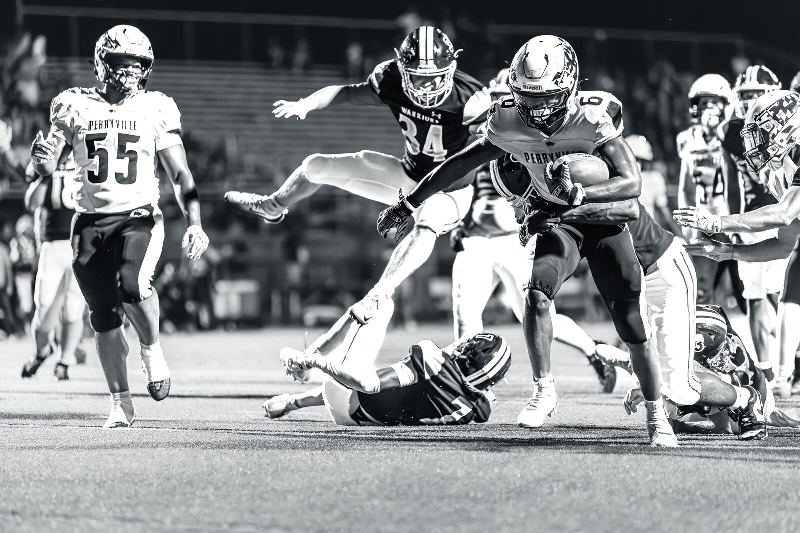 Black and white photo of a running back leaping over a fallen defender during a night game
