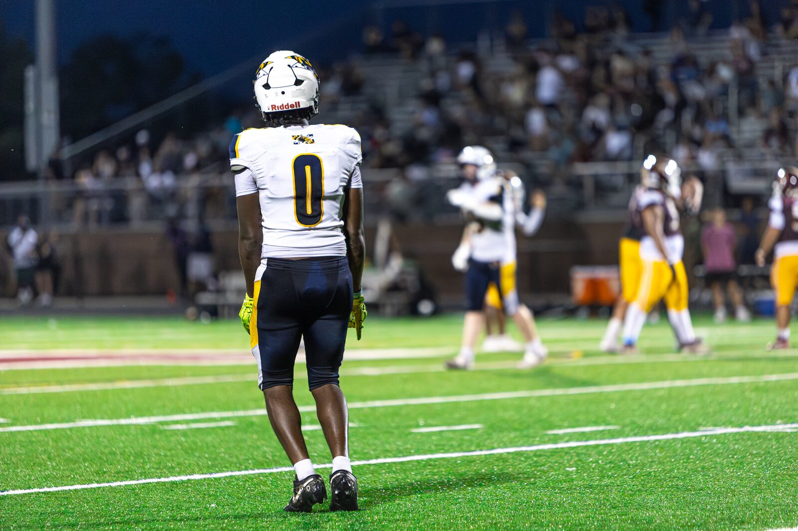 Player number 0 standing alone on the field under the lights while teammates warm up behind him