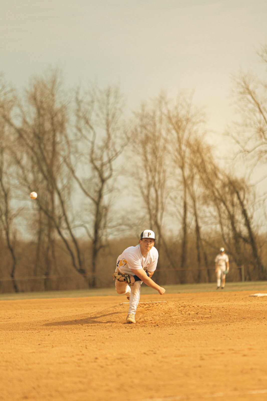 Wide view of a pitcher on the mound in golden light with the ball in the air and bare trees beyond the outfield